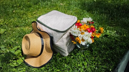 Straw hat, cooler bag and colorful bouquet of wildflowers on green grass in sunny park. Concept of summer picnic, outdoor leisure and enjoying peaceful nature moments
