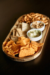 Wooden platter with assorted snacks: chips, crackers, pretzels, dip, nuts displayed on table