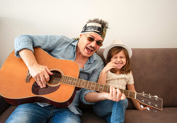 father and daughter laughing together while playing guitar on a sofa at home. A joyful family bonding moment through music and fun. Generation alpha and modern parenting concept.