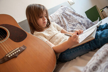 little girl drawing on a sketchbook while relaxing on a bed near an acoustic guitar. A peaceful moment of childhood creativity and imagination at home. Generation alpha concept.