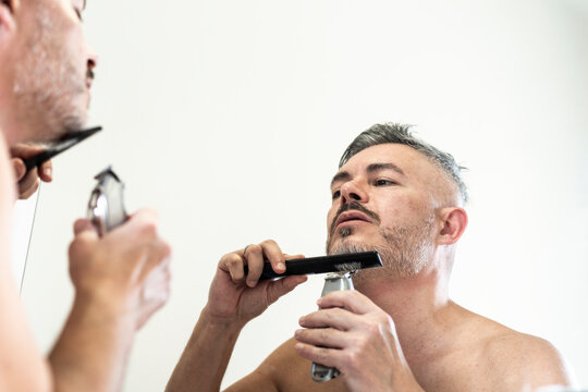 middle-aged man grooming his beard in front of the mirror using an electric trimmer and a comb. The image represents male self-care and daily hygiene. Modern masculinity and personal grooming concept