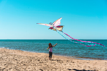 Smiling little girl playing on the beach while flying a colorful butterfly kite over the sea. A joyful and carefree childhood moment by the water. Outdoor activity and summer freedom concept.