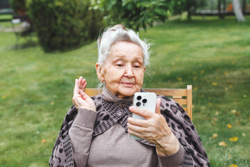 Elderly woman sitting on chair outdoors and waving during video call on smartphone, concept of...