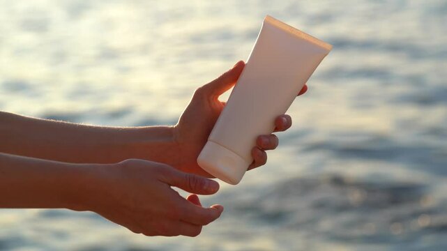 Women's hands opens white mockup tube of cream or lotion and applies to skin, rubbing against background of shiny seawater. The concept of face and body care, sun and uv protection, beauty products