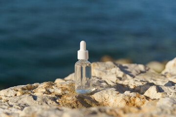 Glass serum bottle with a dropper cap placed on rocky surface with blue ocean in the background....