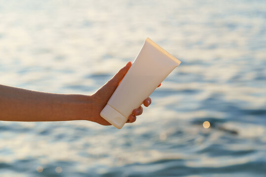 Young woman's hand holds a white mockup tube of cream on a background of sea water illuminated by the setting sun. Concept of beauty products, skin care, sunscreen