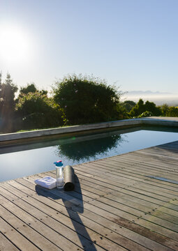 Black yoga mat resting on wooden deck by pool beside white towel and blue-lidded water bottle