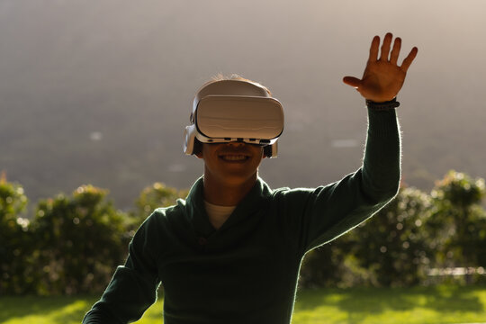 Man is wearing VR headset, raising wristband on grassy lawn near trimmed hedge, with sunlight glow