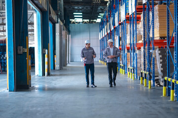 Logistics managers in protective gear walking through a warehouse aisle during an operational discussion. Modern storage system and professional warehouse supervision.