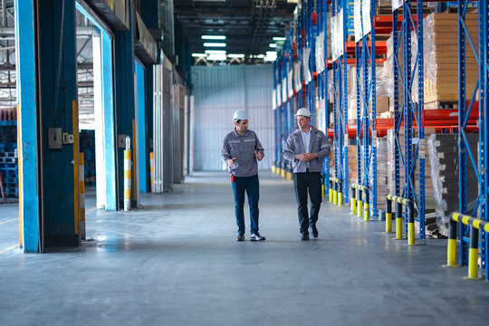 Logistics managers in protective gear walking through a warehouse aisle during an operational discussion. Modern storage system and professional warehouse supervision.