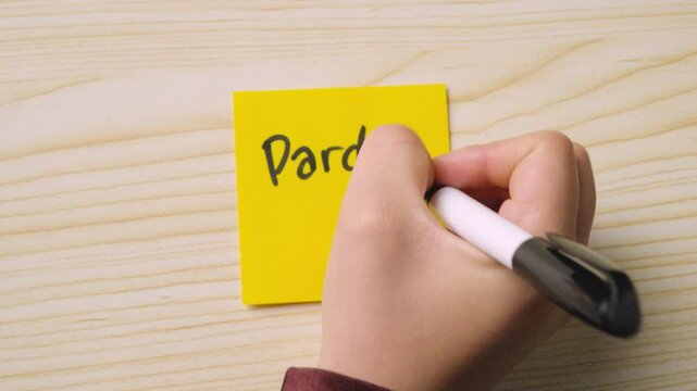 A hand writes &ldquo;Pardon me&rdquo; with a sad face on a yellow sticky note placed on a light wood surface, conveying an apology.