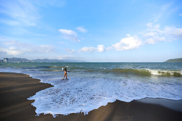A woman in a bikini stands amidst frothy ocean waves breaking on a wide sandy beach, enjoying the...