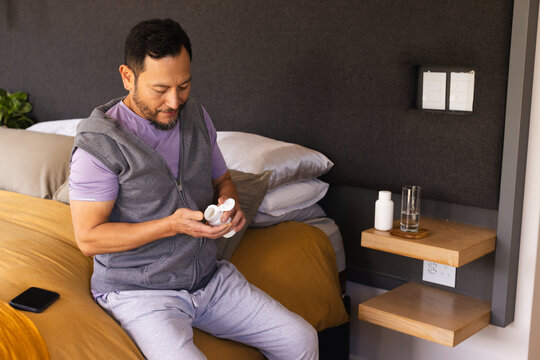 Asian man holding white pill bottle and inspecting pill on bed with smartphone in bedroom