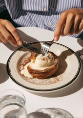 Close-up hands cutting warm pastry topped with melting vanilla ice cream and cocoa powder on white plate, cozy dessert moment in sunlight, striped shirt, elegant brunch atmosphere.
