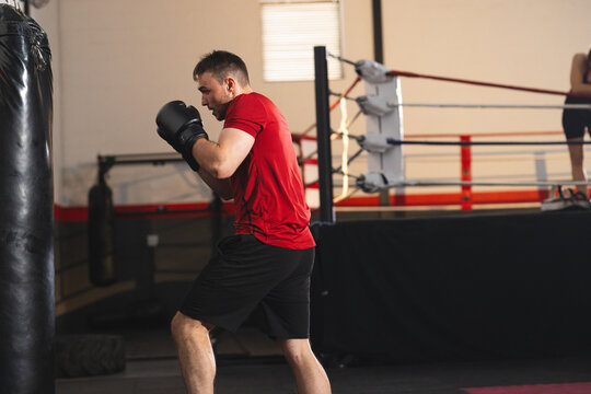 Boxer wearing red shirt, black gloves practicing jabs on heavy bag near boxing ring in gym