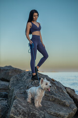 beautiful brunette woman wearing fitness attire stands on a rock by the beach She enjoys a serene sunset stroll with her white dog beside her, showcasing her smooth skin after epilation