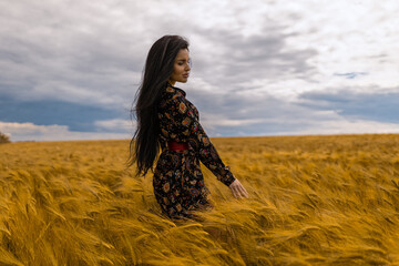 brunette woman in a floral dress stands gracefully in a golden wheat field, her hair gently swaying...