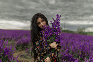 beautiful brunette stands in a vibrant field of delphiniums, holding a bouquet of purple flowers...