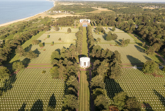 Aerial view of rows of white crosses stretching across the green landscape towards the sea, with white monuments standing in solemn remembrance, Colleville-sur-Mer, Normandie, France.