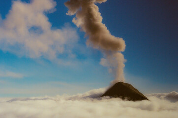 A stunning view of Fuego Volcano erupting with smoke rising into the blue sky above cloud cover. Capturing the raw beauty of nature and the dramatic landscape of a volcano in action. 35mm Film