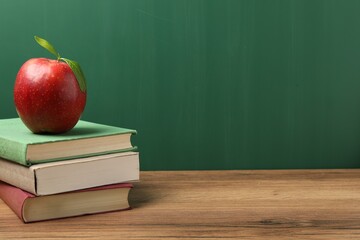 Different books and red apple on wooden table near blackboard, closeup. Space for text