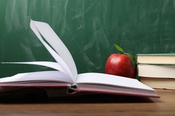 Different books and red apple on wooden table near blackboard, closeup