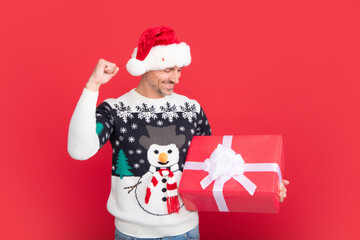 Santa with christmas gift box. Studio portrait of guy wears santa hat and winter sweater. Man with christmas hat over isolated red background.