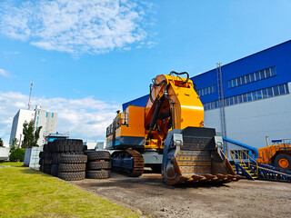 Large orange excavator is parked on construction site, surrounded by stacked tires and industrial buildings, showcasing heavy machinery in action