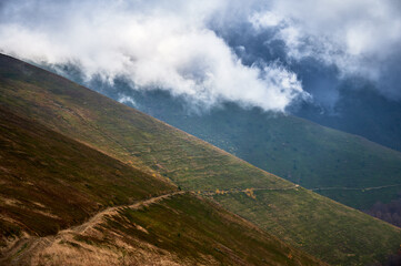 Steep mountainside covered in lush green grass and patches of brown vegetation, with winding dirt...