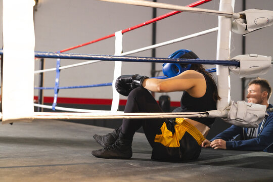 Female boxer in headgear sitting in boxing ring corner, trainer holding towel by ropes, copy space