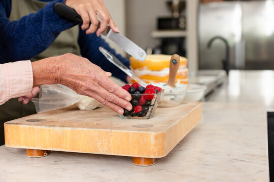 Senior female friends assembling cake with raspberries in home kitchen using spatula and frosting