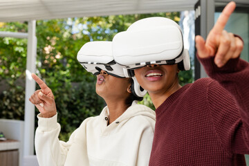 Diverse female friends wearing wireless VR headsets pointing at virtual interface under pergola