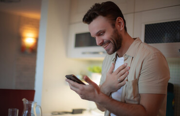 Smiling young man in casual clothes looks at his smartphone with hand on chest, feeling touched and...