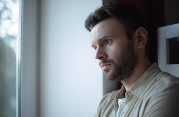 Thoughtful young man in casual clothes looks out the window with a serious and pensive expression,...