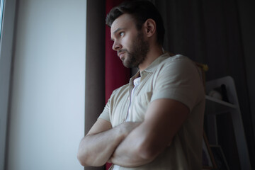 Thoughtful young man in casual clothes stands by a window with arms crossed, looking outside with a...