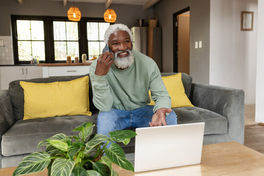 Senior African American man calling on smartphone sitting at home on sofa with laptop on table