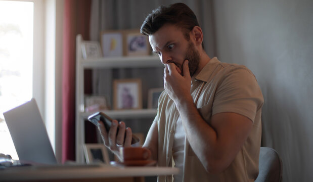 Young man in casual clothes sits at a desk with a laptop, looking at his phone with a shocked, worried expression while covering his mouth indoors.