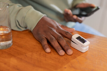 African American male using pulse oximeter, holding smartphone on wooden table with glass of water