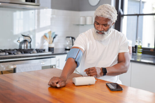 Senior African American man checking blood pressure with digital monitor on modern kitchen island - Powered by Adobe