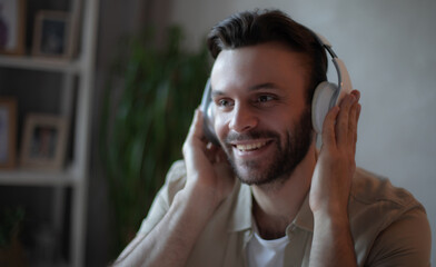 Smiling young man wearing headphones, enjoying music while working from home, sitting at a desk in...
