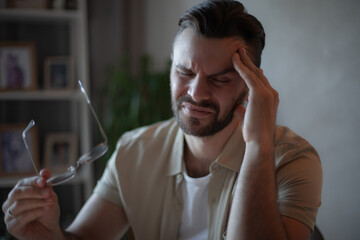 Young man in beige shirt holding glasses and touching his head with a pained expression, suffering...