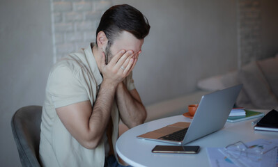 Stressed young man sitting at desk with laptop, covering face with hands in frustration or burnout,...