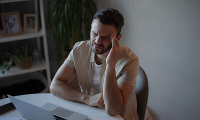 Frustrated young man sitting at desk with laptop, holding his head in pain with eyes closed,...