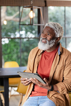 Senior African American man sitting at table holding tablet under metal pendant light near window