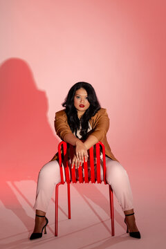 Asian woman posing backwards on red metal chair in studio, wearing tan blazer and pink trousers