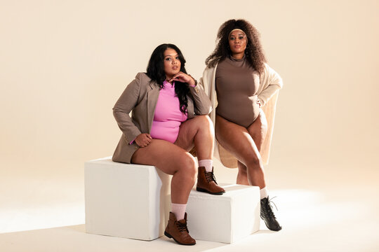 Diverse female friends posing on white blocks in cream studio wearing bright bodysuits and boots