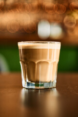 Clear glass of creamy coffee with foamy top on dark wooden table in cozy cafe setting