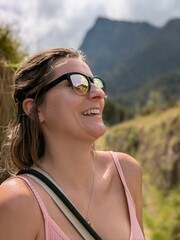 Happy blond teenage girl wearing sunglasses, smiling and walking on a sunny mountain trip; playful, fit and expressive young woman enjoying warmth, wellness, and a joyful hiking journey
