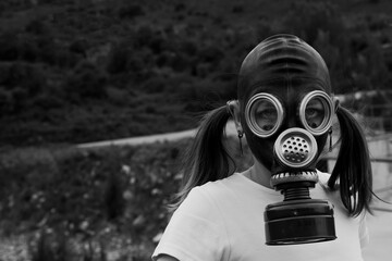 A young woman with pigtails wears a gas mask while standing outdoors in a natural environment