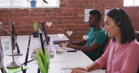 Middle Eastern woman in pink blouse in headset typing in office, monitors potted plants, copy space